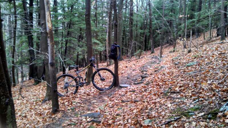 A mountain bike resting on a trail in a forested area, surrounded by trees and a ground covered with autumn leaves. A backpack is hanging from a tree nearby, with rocky terrain visible along the path. Kingman Farm mountain bike trail.