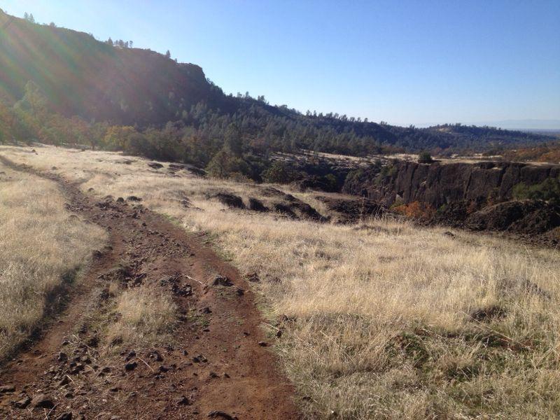 A dirt trail winding through dry grassland, leading toward a rocky terrain and distant hills under a clear blue sky. Sunlight creates a bright lens flare on the left side of the image. Upper Bidwell Park mountain bike trail.
