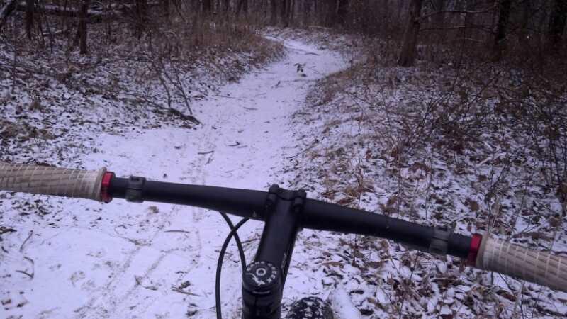 Mountain bike handlebars visible in the foreground, with a snowy trail extending through a wooded area in the background. The path is surrounded by grass and leaf litter, indicating winter conditions. Rock Bridge Memorial State Park mountain bike trail.