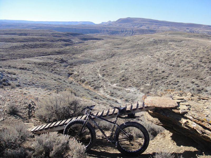 A mountain bike is resting on a wooden ramp overlooking a vast, arid landscape with rolling hills and a winding dirt path in the distance. The scene captures a clear blue sky and the rugged terrain typical of desert-like environments. Wilkins Peak Trails mountain bike trail.