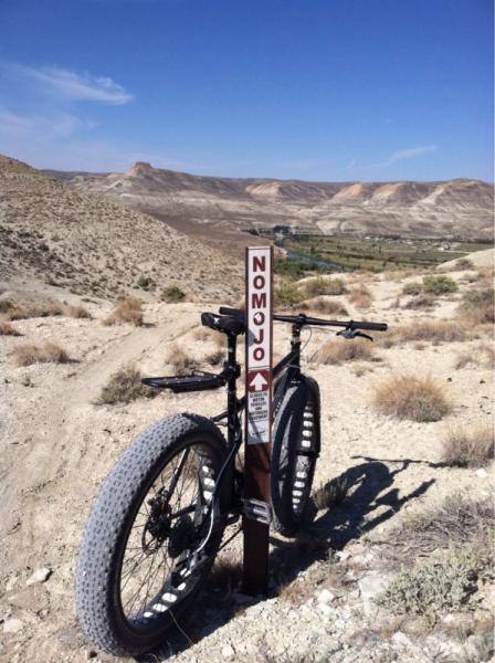 A fat bike parked next to a trail sign pointing to "Nom Jo," set against a backdrop of rugged hills and a clear blue sky. The landscape features dry terrain with sparse vegetation, showcasing a remote outdoor environment ideal for biking. NoMoJo mountain bike trail.