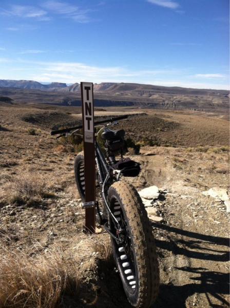 A mountain bike parked beside a trail sign labeled "TNT," with a vast landscape of rolling hills and mountains in the background under a clear blue sky. The foreground features dry grass and rocky terrain typical of a rugged outdoor setting. Tnt mountain bike trail.