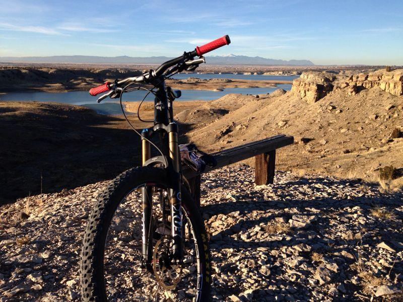 A mountain bike with red handlebars is positioned on rocky terrain, overlooking a serene landscape of hills and a winding body of water in the distance. The sky is clear with soft daylight, highlighting the beauty of the outdoor scenery. A wooden bench is nearby, adding to the peaceful setting. South Shore Lake Pueblo mountain bike trail.