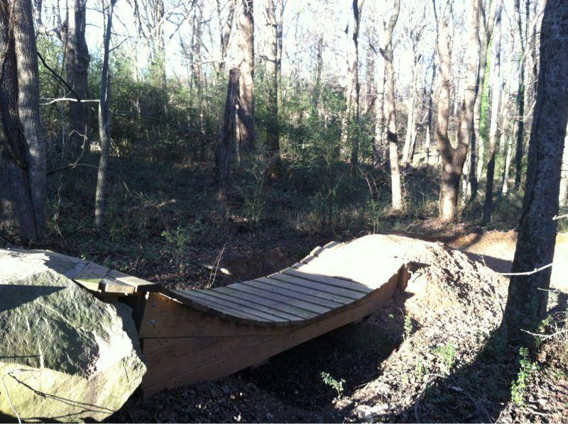 A wooden bridge crossing over a small gap in a wooded area, surrounded by trees and underbrush. Sunlight filters through the branches, casting shadows on the bridge and the ground. Back Yard Trails mountain bike trail.