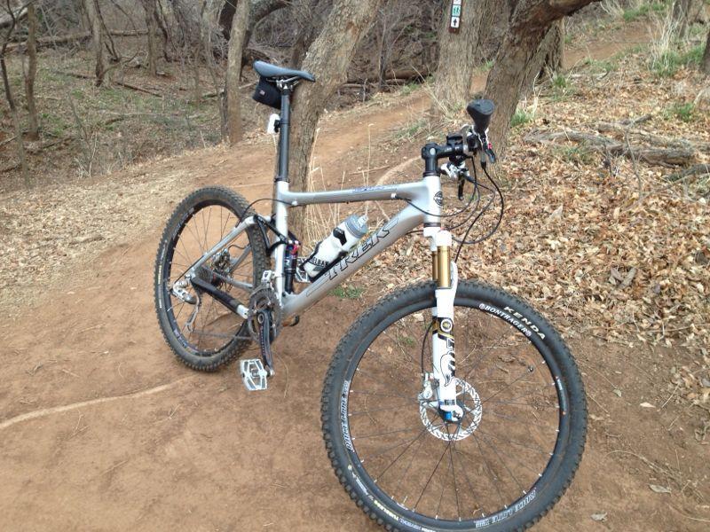 A silver mountain bike positioned on a dirt trail surrounded by trees and dry leaves. The bike features front suspension, a water bottle mounted on the frame, and is parked beside a natural pathway. Bluff Creek Trail mountain bike trail.