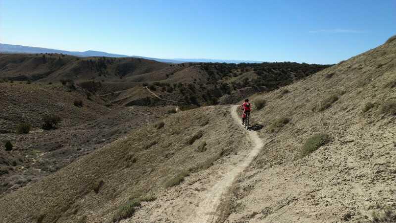 A mountain biker riding along a narrow dirt trail on a hillside, surrounded by rolling hills and sparse vegetation under a clear blue sky. 18 Road Trails / North Fruita Desert mountain bike trail.