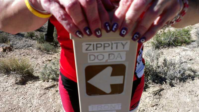 A close-up image of a person holding a directional sign labeled "ZIPPITY DO DA" with an arrow pointing left. The person's nails are elaborately painted, and they are wearing a red shirt. The background features a natural landscape with sparse vegetation. Zippety Do Dah mountain bike trail.