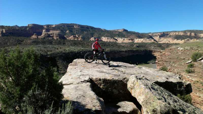 A child wearing a red and white jersey stands on a large rock outcrop with a mountain bike beside them, overlooking a scenic landscape of cliffs and a river below. The sky is clear and blue, enhancing the vibrant colors of the natural surroundings. Kokopelli Trail mountain bike trail.