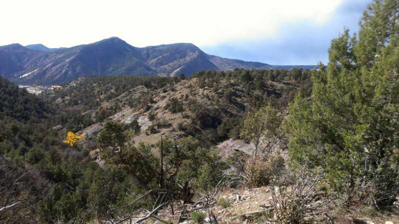 A scenic view of rolling hills and mountains under a partly cloudy sky, with patches of green vegetation and scattered trees in the foreground. The background features towering mountains with varying shades of green and brown, indicating a natural landscape. Horse Gulch mountain bike trail.