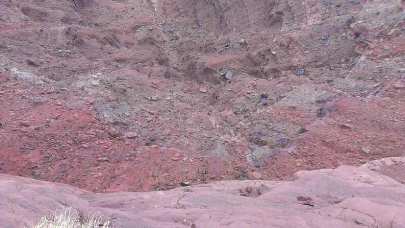 Aerial view of a steep, rocky canyon with reddish-brown soil and scattered rocks, showcasing layers of sediment and erosion patterns. Dead Horse Point State Park mountain bike trail.