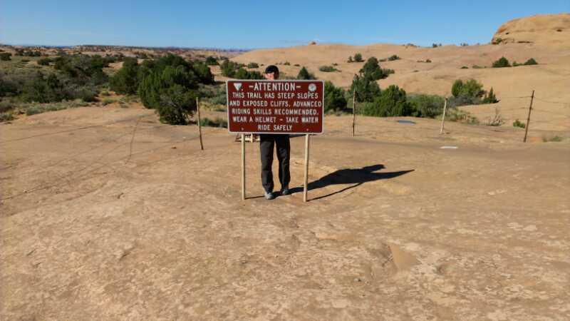 A person stands next to a warning sign on a rocky trail, surrounded by a desert landscape. The sign indicates that the trail has steep slopes and exposed cliffs, advising riders to wear helmets, stay hydrated, and have advanced riding skills. Slickrock mountain bike trail.
