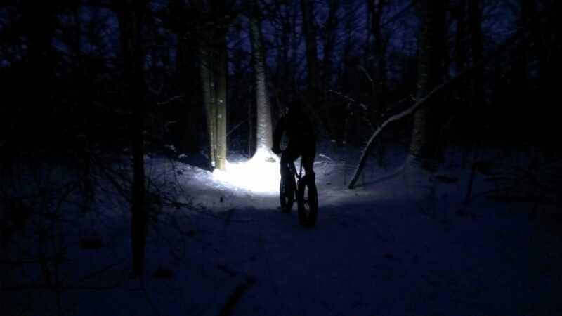 A person riding a fat bike through a snowy forest at night, illuminated by the beam of a bright headlight, with dark trees surrounding them. Kettle Moraine John Muir + Emma Carlin mountain bike trail.