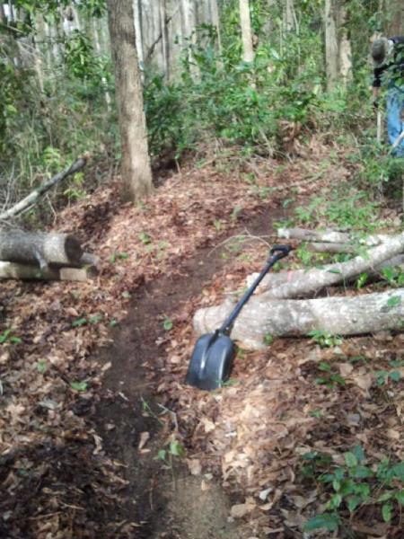 A narrow dirt path through a wooded area, with a black shovel resting on the ground amid scattered leaves and tree branches. Logs are visible along the side of the path, surrounded by dense greenery. Boyd Pond mountain bike trail.
