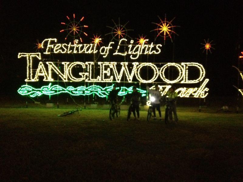 Brightly lit sign reading "Festival of Lights Tanglewood Park" against a night sky, with decorative starburst lights surrounding it. Silhouettes of people stand in front, some with bicycles, creating a festive atmosphere. Tanglewood Park mountain bike trail.
