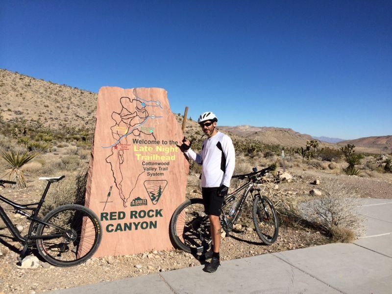 A cyclist in a white long-sleeve shirt and helmet stands next to a large rock sign that reads "Welcome to the Late Night Trailhead, Cottonwood Valley Trail System, Red Rock Canyon." Two mountain bikes are parked beside the sign, with arid desert landscape and mountains in the background under a clear blue sky. Blue Diamond mountain bike trail.
