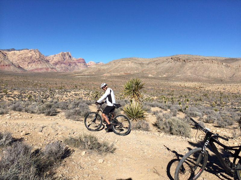A mountain biker in a white long-sleeve shirt and helmet, pausing on a dirt trail surrounded by rugged desert terrain and mountains under a clear blue sky. A second bike is partially visible in the foreground. Small desert plants and shrubs dot the landscape. Blue Diamond mountain bike trail.