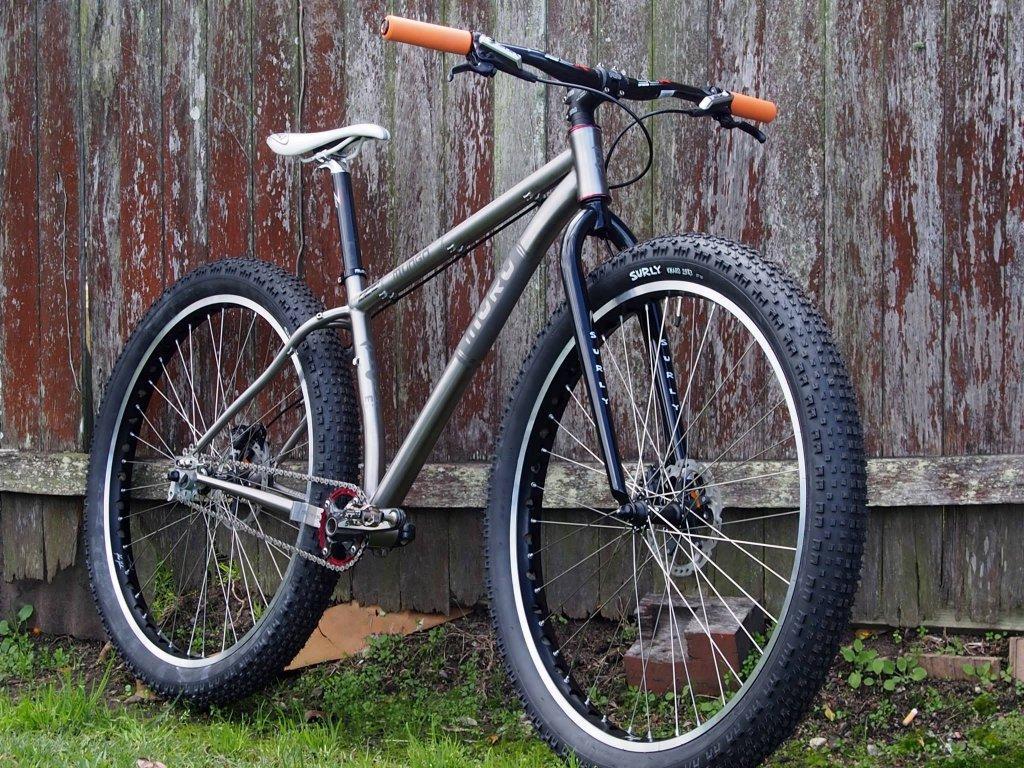 Muru Mungo 29er plus: A silver and black mountain bike with thick tires and orange handlebars, leaning against a weathered wooden fence. The bike features a sleek frame, disc brakes, and a single-speed setup, ready for outdoor adventures.