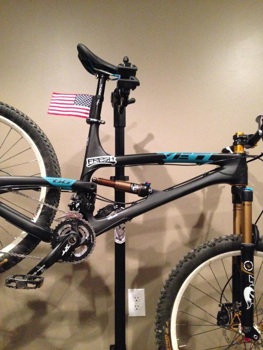 Yeti SB66c: A mountain bike with a sleek black frame featuring turquoise accents is mounted on a bike stand. The bike has a small American flag attached near the seat post. It showcases a well-maintained drivetrain with visible gears and a sturdy set of tires. The background is a neutral-toned wall, and the bike stand is positioned near an electrical outlet.