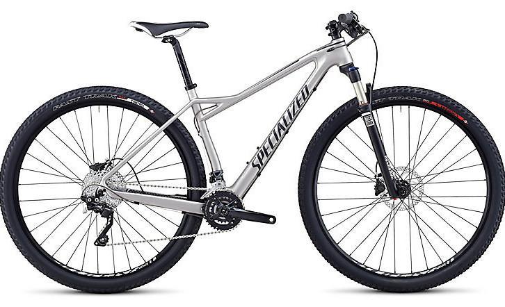 Specialized Fate Comp Carbon 29: A silver mountain bike with oversized tires, featuring a front suspension fork, disc brakes, and a lightweight aluminum frame. The bike has a sleek design with the brand name "Specialized" prominently displayed on the frame.