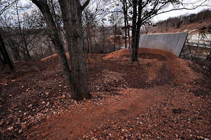 A winding dirt bike trail surrounded by trees, leading to a large wooden ramp. The ground is covered in brown dirt and scattered leaves, with a sloped incline in the background. The area appears quiet and natural, with sparse foliage overhead. Slaughter Pen Trail mountain bike trail.
