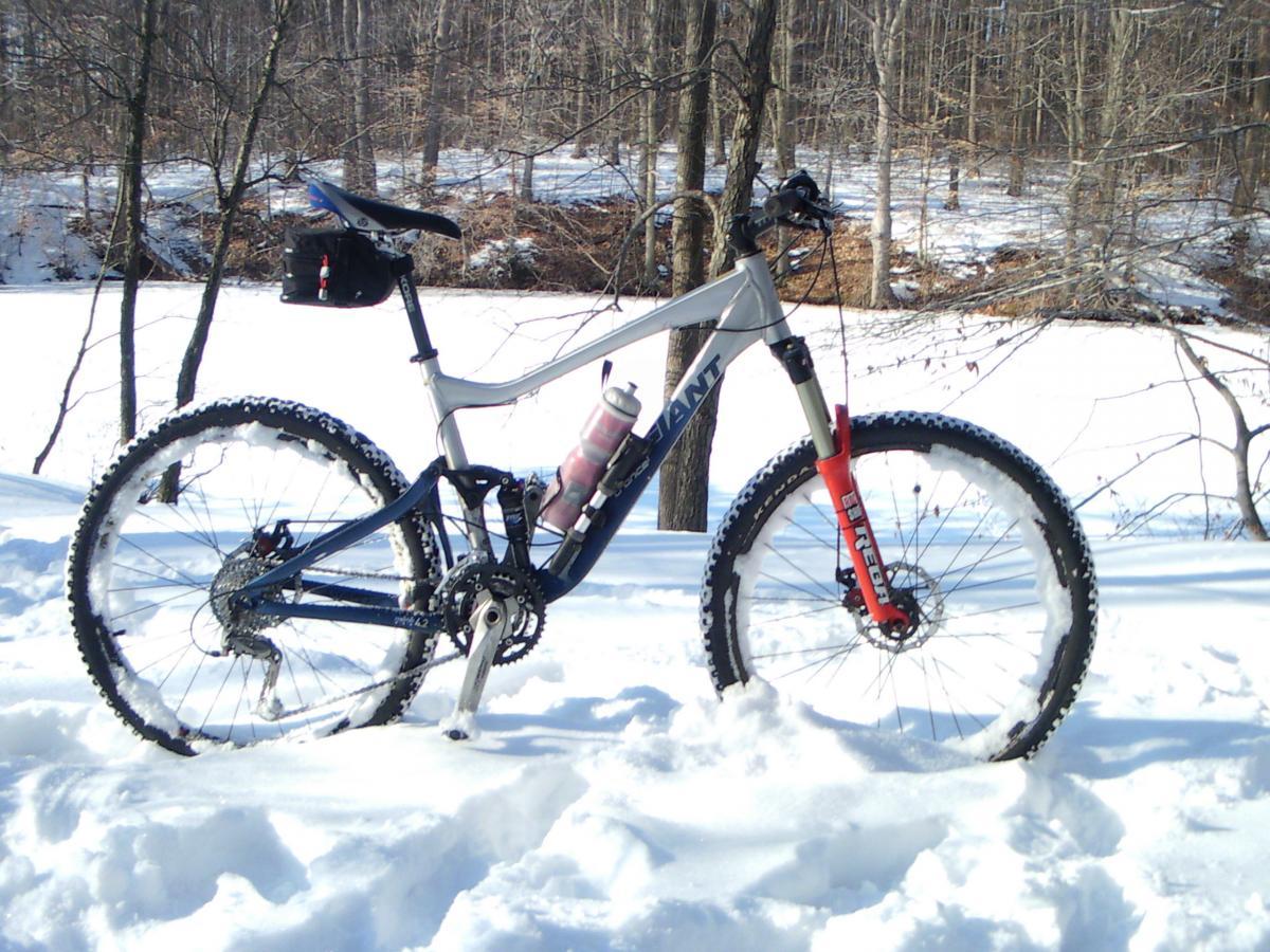 Giant Trance X1: A mountain bike with a blue and gray frame standing in a snowy landscape, featuring snow-covered wheels and a black backpack attached to the seat. In the background, a serene winter scene with bare trees and a frozen pond.