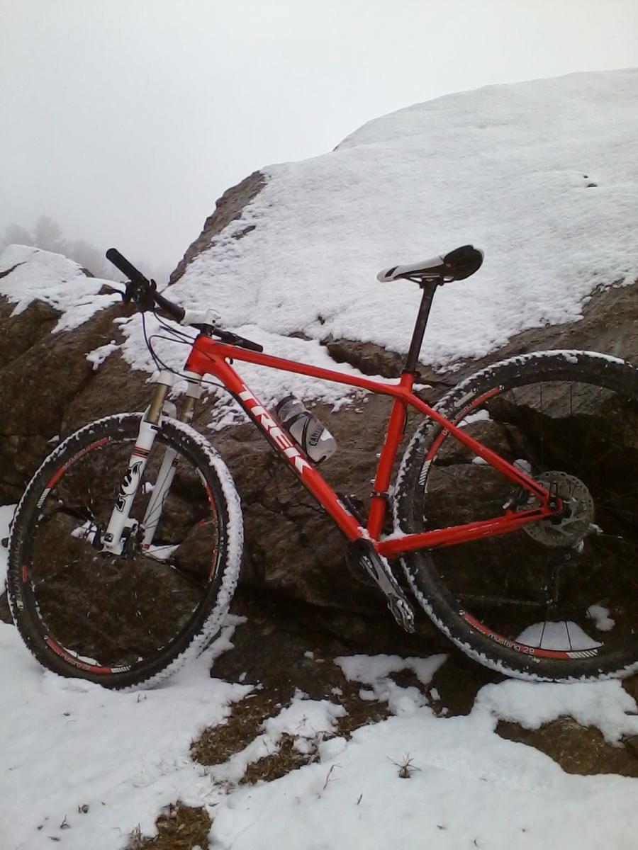 Trek Superfly 8: A bright red mountain bike with a sturdy frame and wide tires resting against a rock, partially covered in snow. The background features a snowy landscape with a foggy sky.