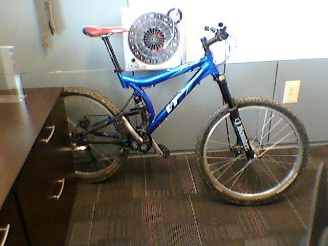 Giant VT2: A blue mountain bike with a red seat is leaning against a grey wall in an office setting. The bike has visible dirt on its tires, indicating recent outdoor use. In the background, there is a mechanical fan mounted on the wall and a wooden cabinet with drawers. The flooring is carpeted with a geometric pattern.