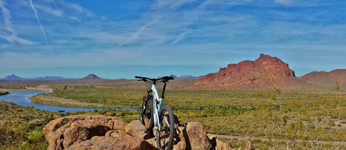 A mountain bike parked on a rocky outcrop overlooking a scenic landscape with a winding river, lush greenery, and distant mountains under a clear blue sky. Hawes Loop mountain bike trail.