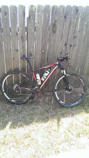 Specialized Epic Comp 29er: A mountain bike with a black and red frame, two water bottle holders, and knobby tires, leaning against a wooden fence. The ground is grassy and the image is taken in daylight.