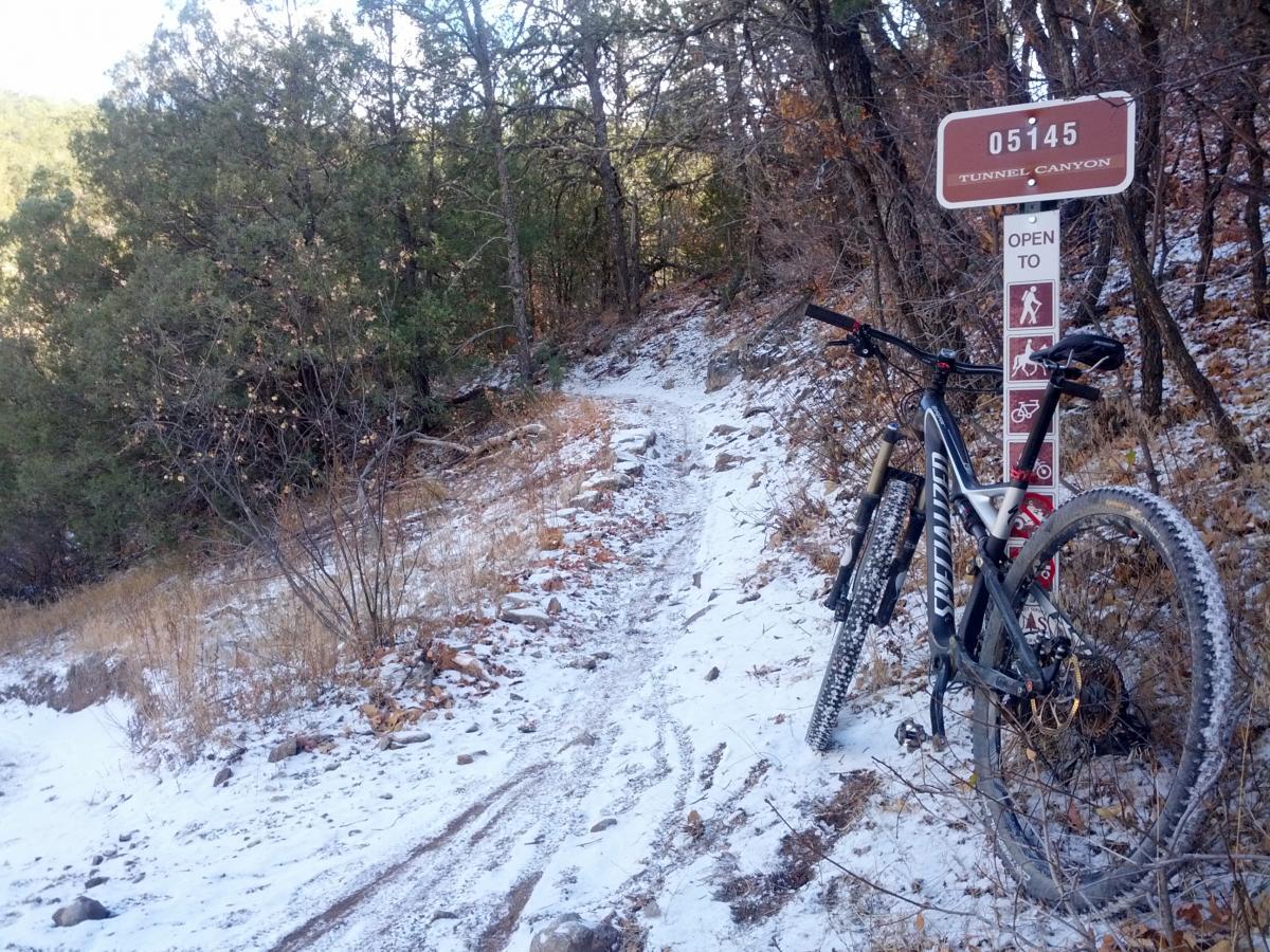 A mountain bike leans against a trail sign that reads "05145 Tunnel Canyon" in a snowy outdoor landscape. The path, partially covered in snow, winds through trees and dry vegetation, indicating it is open to various activities. The scene captures the essence of a winter biking adventure in a wooded area. Tunnel Canyon mountain bike trail.