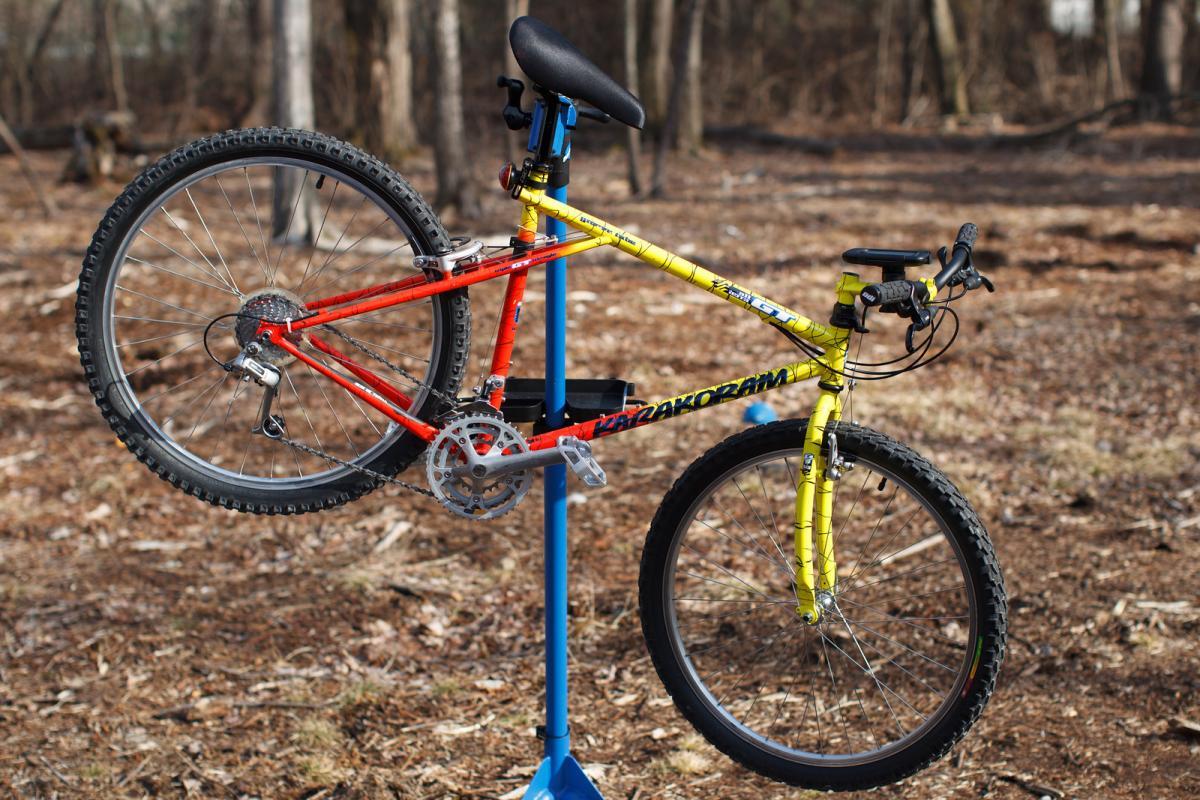 GT Karakoram: A brightly colored mountain bike is positioned on a blue repair stand in a wooded area. The bike features a mix of red and yellow frames, showcasing its distinct design. The background consists of a natural setting with dry leaves and trees, indicating an outdoor environment suitable for biking.