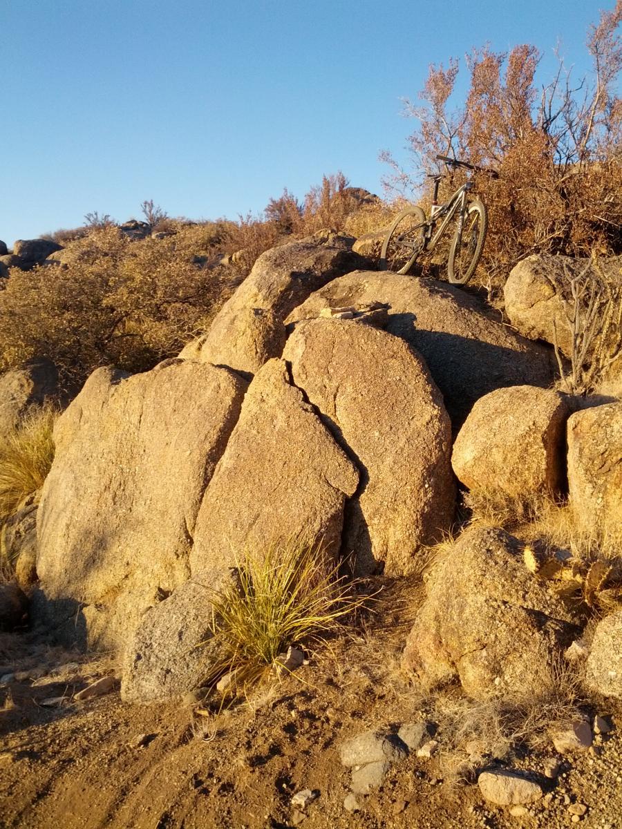 A mountain bike resting on a large rock formation surrounded by dry vegetation and sparse grass under a clear blue sky. South Foothills mountain bike trail.