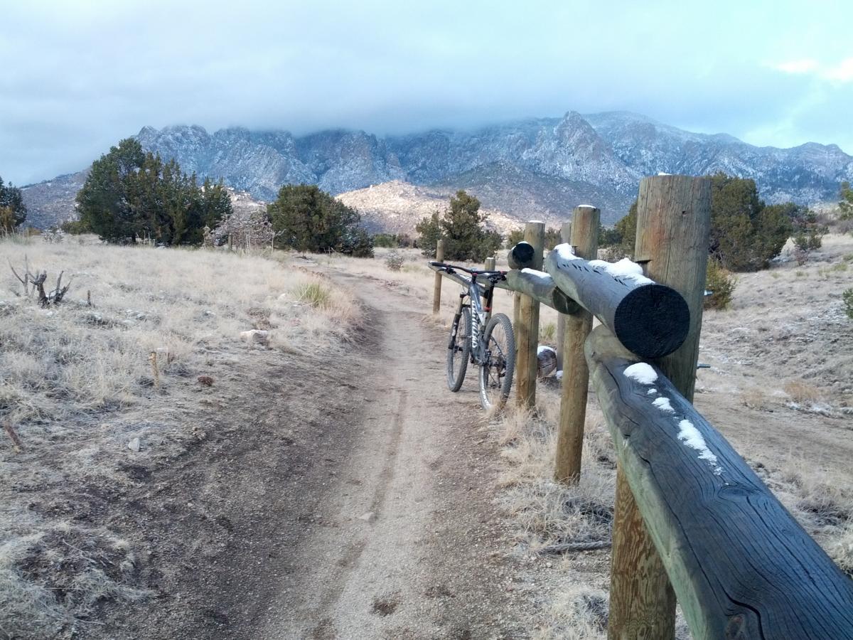 A mountain bike leaning against a wooden fence on a dirt trail surrounded by dry grass and sparse greenery, with snow-capped mountains in the background under a cloudy sky. Elena Gallegos Open Space / North Foothills mountain bike trail.