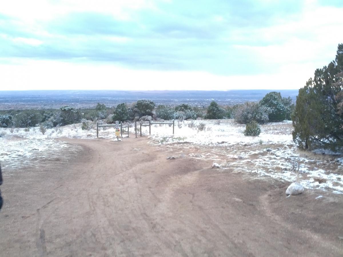 A winding dirt path leads to a wooden gate in a snow-dusted landscape, with patches of vegetation on either side. In the background, a vast view of distant hills and a cloudy sky stretches out over the horizon, creating a serene winter scene. Elena Gallegos Open Space / North Foothills mountain bike trail.