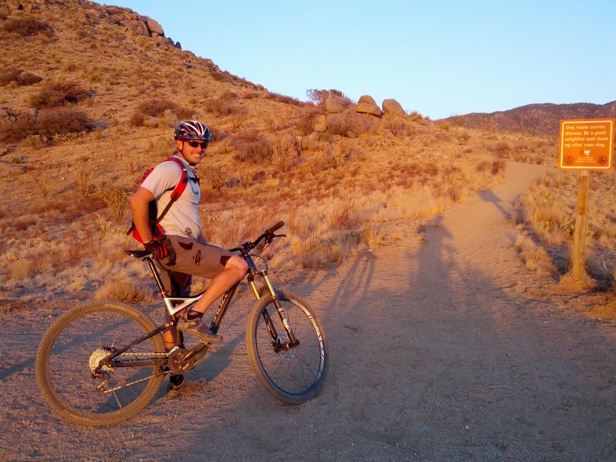 A mountain biker in a helmet and sunglasses poses with his bike on a dirt trail surrounded by arid landscape and a sign warning about dog waste. The scene is illuminated by warm, golden sunset light, highlighting the rocky terrain and sparse vegetation. South Foothills mountain bike trail.