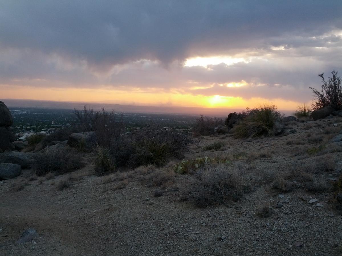 A scenic view from a rocky hillside at sunset, showcasing a dramatic sky filled with clouds and shades of orange and yellow. In the foreground, dry shrubs and grasses are visible on the rugged terrain, with distant hills and a cityscape faintly visible below. South Foothills mountain bike trail.