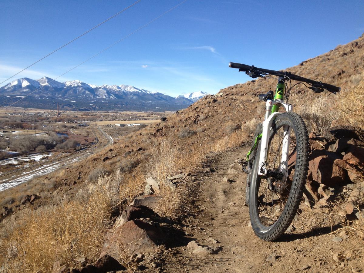 A mountain bike rests on a dirt trail with rocky terrain, overlooking a valley and distant snow-capped mountains under a clear blue sky. Brown grass and sparse vegetation surround the bike, highlighting the natural landscape. Arkansas Hills mountain bike trail.