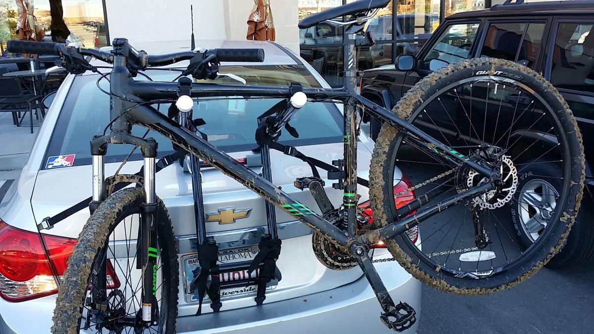 Giant Talon 27.5: A black mountain bike, covered in mud, is mounted on a bike rack attached to the back of a silver Chevrolet car. The bike's front wheel is secured, with a close view of the handlebars and frame. The background features other parked cars and a patio area.