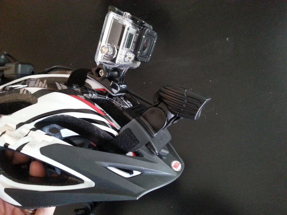 GoPro Hero3: A close-up image of a bicycle helmet featuring a mounted action camera. The helmet is predominantly white with black and red accents. The camera is securely attached to the top of the helmet with a black mounting clip, ready for capturing video footage. The background is a solid black surface.