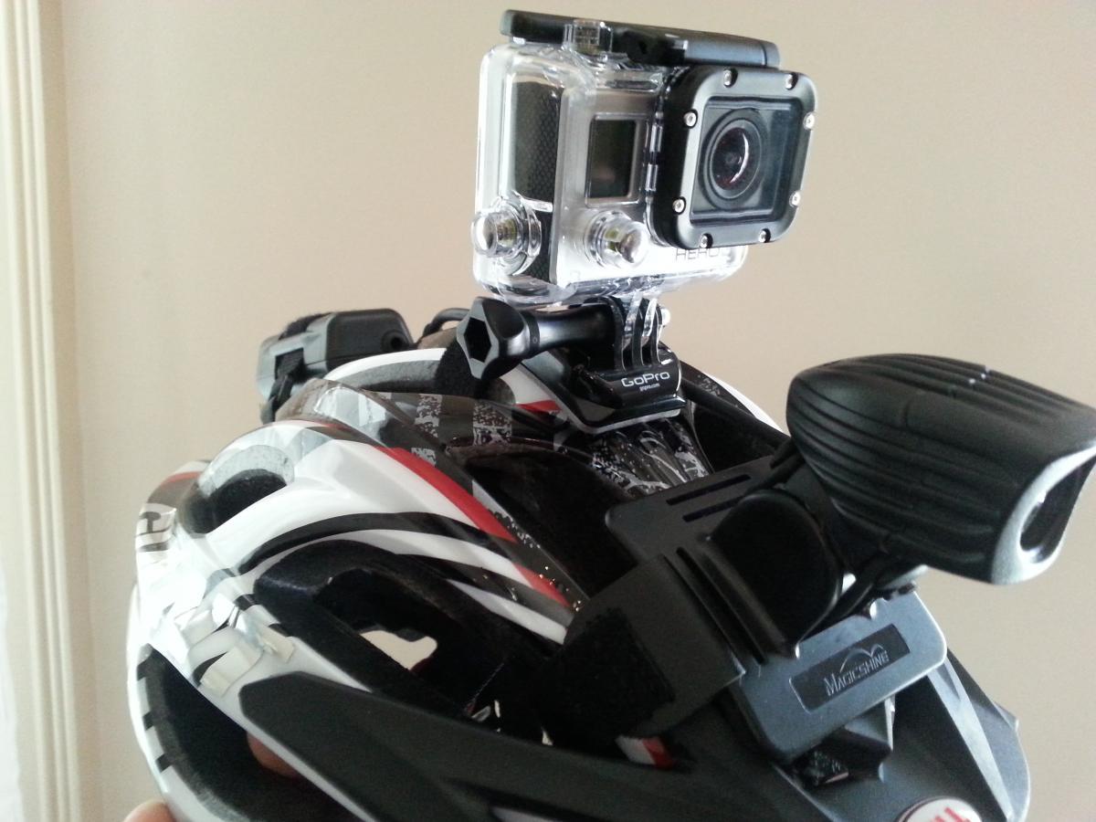 GoPro Hero3: A close-up image of a bicycle helmet equipped with a mounted action camera and a front light. The helmet features a white and black design with red accents. The action camera is securely attached to the top of the helmet, showcasing its compact and rugged design. The background is neutral, emphasizing the helmet and its accessories.