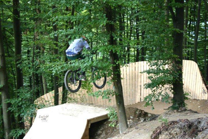 A cyclist performing a jump on a mountain bike over a wooden ramp in a forested area, surrounded by trees and green foliage. Bikepark Winterberg mountain bike trail.