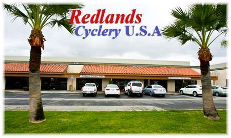 A storefront view of Redlands Cyclery U.S.A., featuring a landscaped area with palm trees in the foreground and several parked cars in front of the building, which has a tiled roof and large windows. The name "Redlands Cyclery U.S.A." is prominently displayed in bold, colorful lettering.