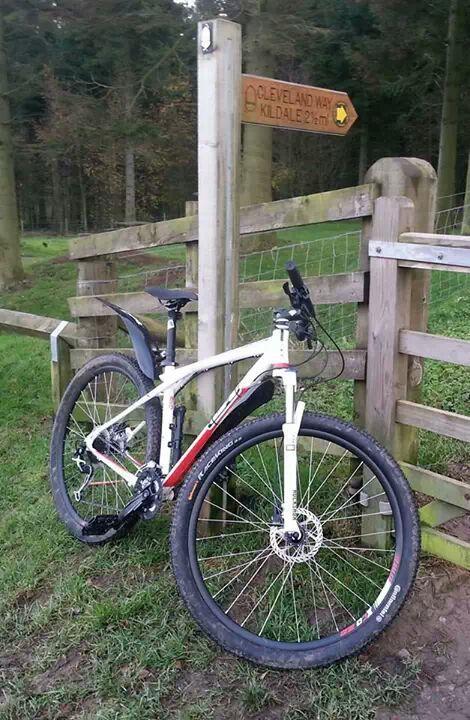 A mountain bike leaning against a wooden gate with a directional sign indicating "Cleveland Way" and "Kildale 2.7 miles" in a forested area.