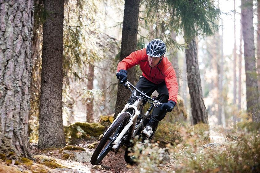 A mountain biker wearing a red jacket and a helmet maneuvers sharply along a dirt trail in a wooded area, surrounded by trees and underbrush. The rider leans into the turn, demonstrating skill and agility on the bike.