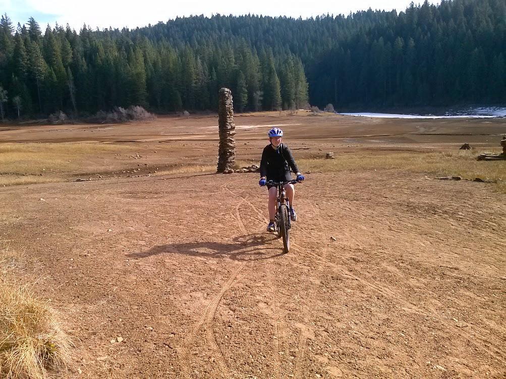 A person riding a mountain bike on a dirt path in a dry landscape, with a backdrop of coniferous trees and a distant body of water. The scene is set in a natural environment, showcasing the rugged terrain and clear skies. Sly Park mountain bike trail.