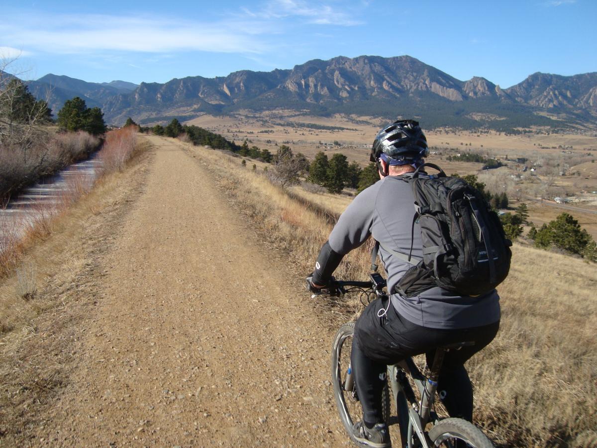A person riding a mountain bike along a dirt path, surrounded by a scenic landscape featuring mountains and valleys under a clear blue sky. Community Ditch mountain bike trail.