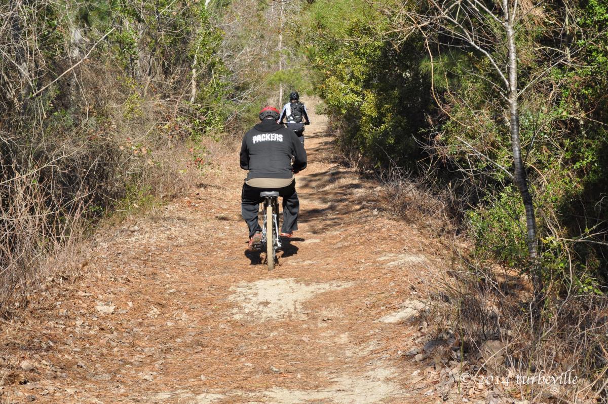 Two cyclists riding along a dirt path surrounded by greenery and trees. The first cyclist, wearing a black jacket with "PACKERS" on the back, is in the foreground, while a second cyclist follows in the background. The trail is lined with dried leaves and pine needles, creating a natural setting for outdoor biking. Horry County Bike Run Park mountain bike trail.