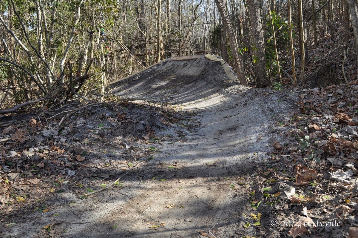 A winding dirt trail surrounded by trees and underbrush, with a gently sloped bank on one side. The ground is covered in fallen leaves and soil, suggesting a natural setting suitable for outdoor activities. Horry County Bike Run Park mountain bike trail.