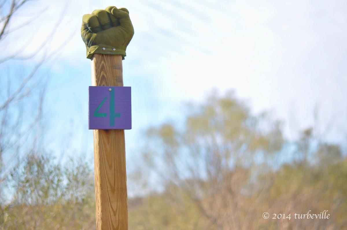 A wooden post displaying a purple sign with the number "4," topped with a green gloved fist. The background features blurred foliage and a clear blue sky, creating an outdoor setting. Horry County Bike Run Park mountain bike trail.
