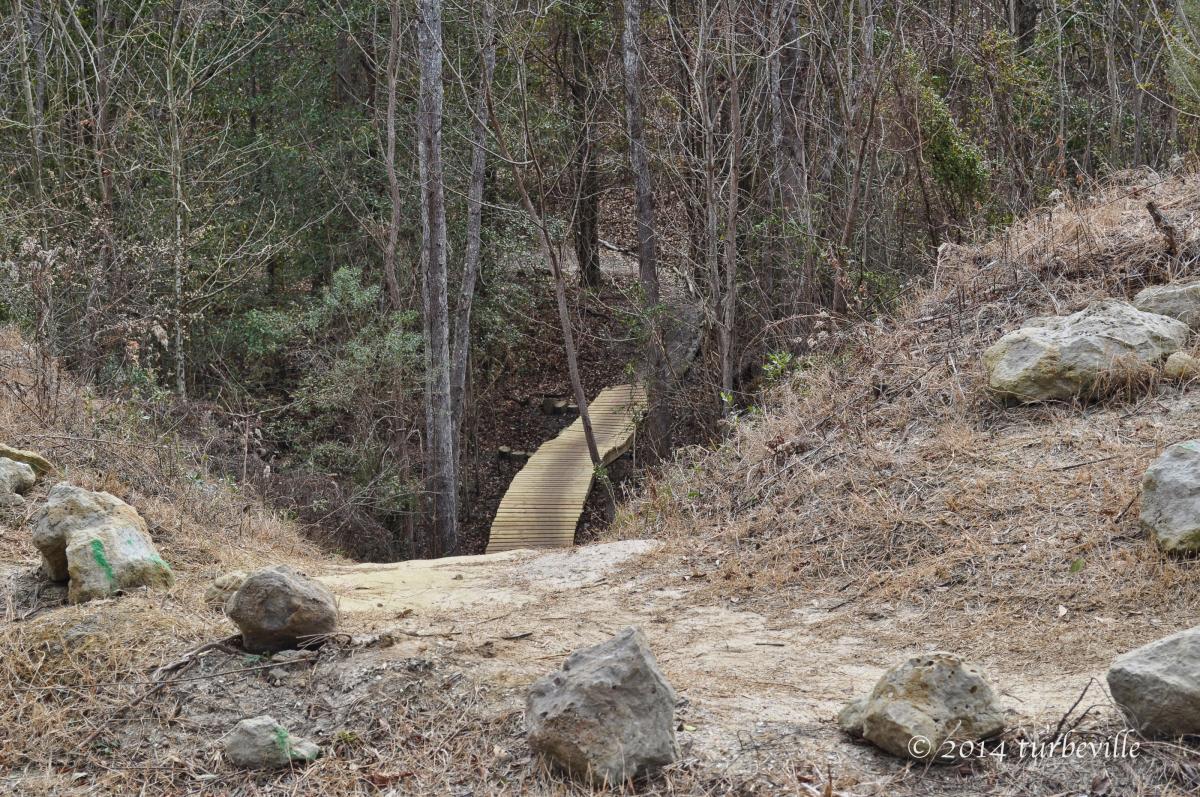 A winding wooden boardwalk leads through a clearing in a forest, surrounded by trees and rocky terrain. The path disappears into the foliage, suggesting a tranquil nature trail. The ground is dry with patches of grass, and scattered rocks add texture to the landscape. Horry County Bike Run Park mountain bike trail.